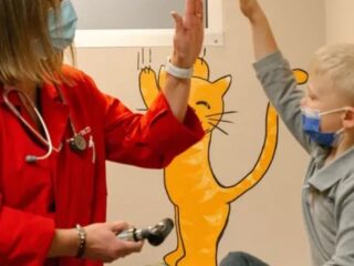 A healthcare worker in a red coat and a young child, both wearing masks, share a high-five. The child sits on an examination table, and there is a painted image of a cat on the wall mimicking the high-five gesture.