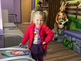 A young girl with pigtails, wearing a red jacket and a colorful skirt, stands smiling in a playful room. A mural with a kangaroo reading to a joey is on the wall behind her. There are chairs and a table with childrens books nearby.